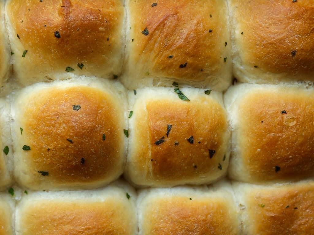 Close-up of freshly baked, golden-brown vegan dinner rolls topped with small flecks of herbs. The rolls are arranged in a grid pattern, showcasing their soft texture and fluffy appearance.